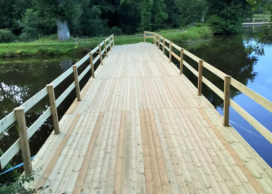 Wooden bridge with handrails crossing over a river, surrounded by greenery and natural landscape
