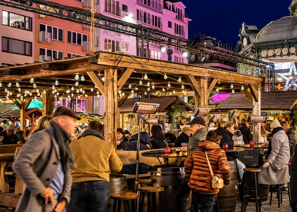 Crowded evening event under a wooden pergola with festoon lighting, bar tables and guests socialising